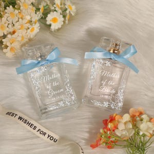 May include: Two clear glass perfume bottles with light blue ribbons, one labeled "Mother of the Groom" and the other "Mother of the Bride." Both bottles have floral designs and the date "18.10.25." The bottles are on a white, fluffy surface with flowers.