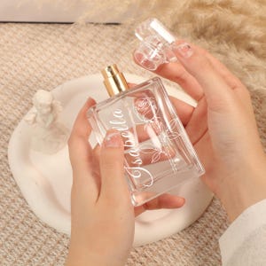 Könnte beinhalten: Eine klare, rechteckige Parfümflasche mit einem goldfarbenen Zerstäuber und einer transparenten Kappe. Die Flasche ist mit einer Rosenillustration und dem Namen "Isabella" in weißer Schrift verziert. Die Flasche wird von einer Person gehalten.