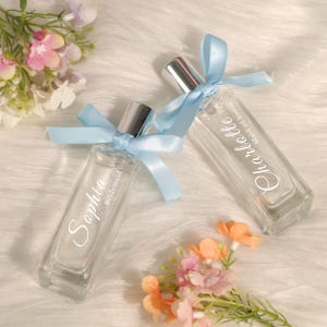 Peut inclure: Deux flacons de parfum en verre transparent avec vaporisateurs argentés, noués de rubans bleu clair. L'un est étiqueté "Sophia, Demoiselle d'honneur", l'autre "Charlotte, Maid of honor". Les flacons sont sur une surface blanche et duveteuse, avec des fleurs en arrière-plan.
