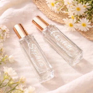 Puede incluir: Dos frascos de perfume rectangulares de vidrio transparente con tapas doradas. Uno está etiquetado "Sophia Bridesmaid" y el otro "Charlotte Maid of honor" en escritura blanca. Los frascos están sobre una superficie blanca, con flores blancas y un sombrero de paja en el fondo.