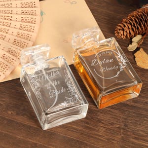 May include: Two clear glass perfume bottles with rectangular shapes and clear stoppers. One bottle is labelled "Mother of the Bride" with floral accents. The other bottle is labelled "Dylan Wendy" with the date "22.05.2025" and floral accents. The bottles are filled with a light amber liquid.