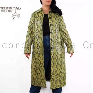 Może przedstawiać: Długi płaszcz zapinany na guziki z wzorem skóry węża w odcieniach zieleni i żółci. Płaszcz jest noszony na czarnym topie i niebieskich dżinsach. Logo marki "SCORPION HIDE CO" jest widoczne w lewym górnym rogu.