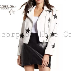 Puede incluir: Chaqueta de cuero blanca con detalles de estrellas negras. La chaqueta tiene un estilo motero clásico con cremallera asimétrica y cuello con solapas. Las mangas y los hombros están adornados con estrellas negras. La marca "SCORPION HIDE CO" es visible.