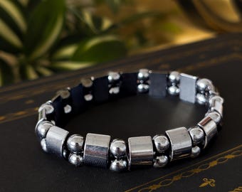 Puede incluir: Una pulsera plateada con una combinación de pequeñas cuentas redondas y cuentas rectangulares más grandes. La pulsera se presenta sobre un fondo oscuro, con un vistazo de vegetación. El diseño es simple y moderno.
