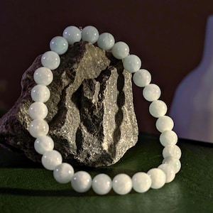 Puede incluir: Un brazalete de cuentas redondas, de color azul pálido y blanco. El brazalete está colocado sobre una piedra gris rugosa, con un fondo oscuro. Las piedras parecen pulidas y tienen un brillo sutil.