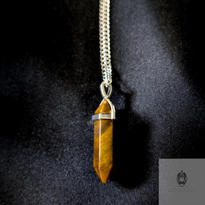 May include: A silver chain necklace with a pendant featuring a polished, elongated, tiger's eye gemstone. The pendant is a warm brown color with dark brown stripes. The necklace hangs against a dark background.