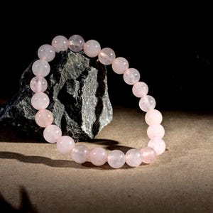 May include: A rose quartz beaded bracelet. The bracelet features round, light pink beads strung together. The beads are smooth and have a subtle, translucent quality. The bracelet is displayed on a neutral surface with a dark background.
