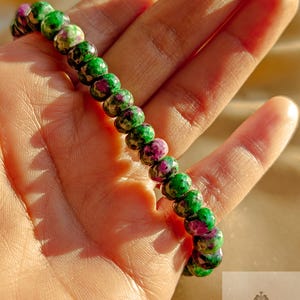 Może przedstawiać: Bransoletka z koralików z naprzemiennymi zielonymi i purpurowymi kamieniami. Bransoletka wykonana jest z małych, okrągłych koralików, tworząc teksturowany i kolorowy dodatek. Bransoletka jest pokazana na dłoni.