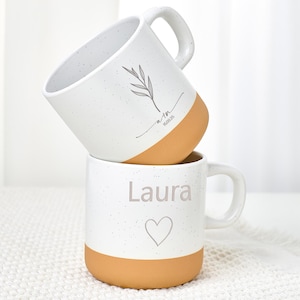 Könnte beinhalten: Zwei gestapelte weiße Keramikbecher mit gesprenkelter Oberfläche und braunen Böden. Der obere Becher hat ein Blattdesign und den Text "a+m 16.08.25". Der untere Becher hat den Namen "Laura" und ein Herzsymbol.