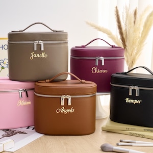 May include: Five personalised cosmetic bags in shades of brown, burgundy, black, pink, and brown. Each bag features a top handle, a silver zip, and a name in gold lettering. Makeup brushes and magazines are in the foreground.