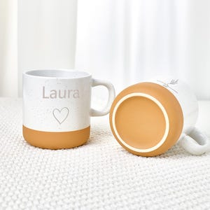 May include: White ceramic mug with a speckled design and a tan base. The mug has the name "Laura" and a heart symbol. Another mug is lying on its side, showing the tan base.