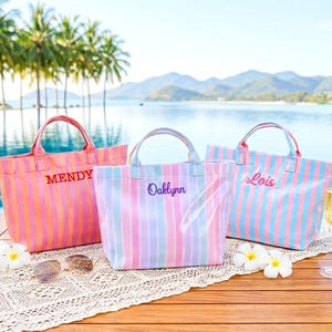 May include: Three rectangular tote bags with vertical stripes and embroidered names. The left bag is orange and pink with MENDY, the center is pink and blue with Oaklynn, and the right is blue and pink with Lois. Each bag has two top handles.