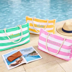 Peut inclure: Trois sacs fourre-tout rectangulaires avec des rayures horizontales blanches et des anses en corde. Un sac est vert avec le nom Dakota, un est jaune avec le nom Maddie, et un est rose avec le nom HANNAH brodé sur le devant. Un chapeau et un livre sont présents.