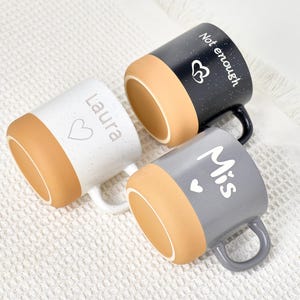 May include: Three ceramic mugs with speckled finishes and tan-colored bases. One mug is white with "Laura" and a heart, another is black with "Not enough" and a heart, and the third is gray with "Mis" and a heart.