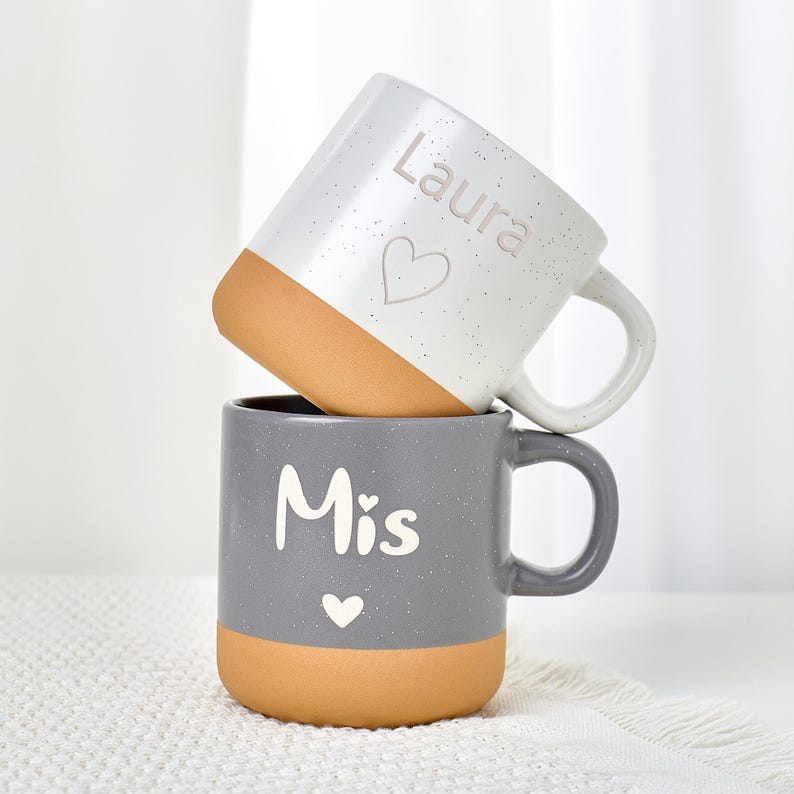 May include: Two ceramic mugs stacked on top of each other. The top mug is white with a brown base and the name "Laura" and a heart. The bottom mug is gray with a brown base and the word "Mis" and a heart. Both mugs have handles.
