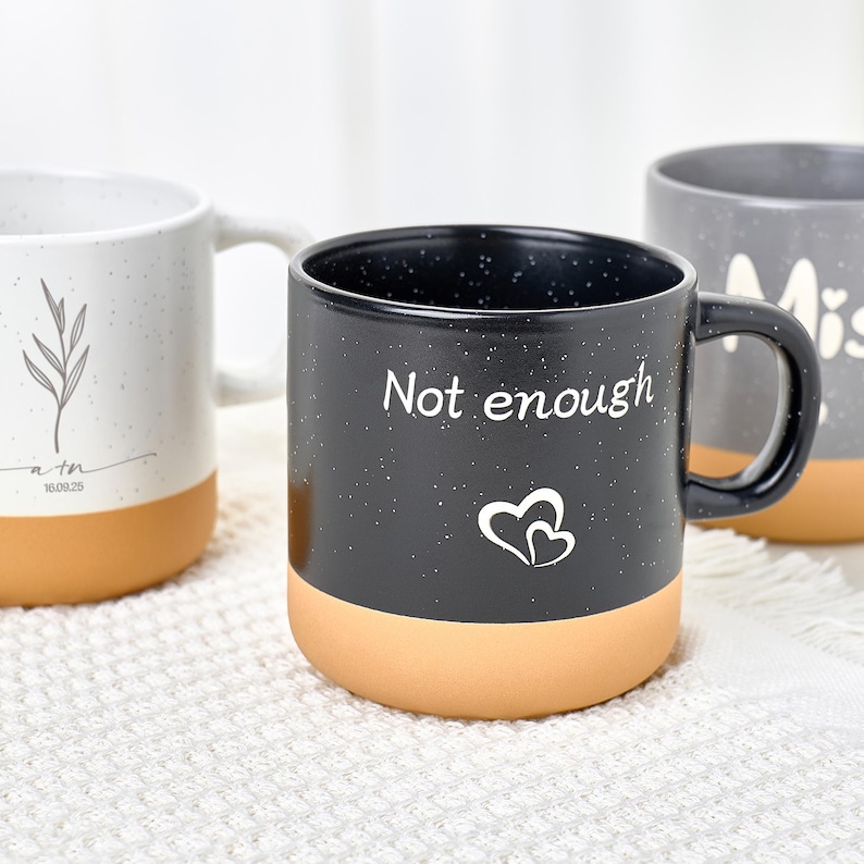 May include: Three ceramic mugs with speckled designs and tan-colored bottoms. The front mug is black with the words "Not enough" and two heart shapes. The other mugs have leaf and word designs.