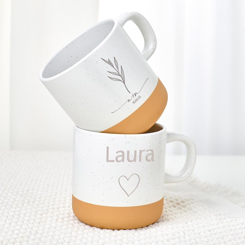 May include: Two stacked white ceramic mugs with speckled finish and tan bottoms. The top mug has a leaf design and the text "a+m 16.08.25". The bottom mug has the name "Laura" and a heart symbol.