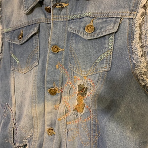 Könnte beinhalten: Eine hellblaue Jeansweste mit einem Used-Look-Design. Die Weste hat goldfarbene Knöpfe, zwei Vordertaschen und dekorative Stickereien mit farbigen Nähten. Die Weste hat einen ausgefransten Rand an den Ärmeln und ein Etikett mit der Aufschrift "tang".