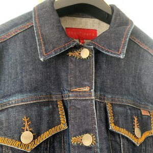 Könnte beinhalten: Dunkelblaue Jeansjacke mit orangefarbenen Nähten und goldfarbenen Knöpfen. Sie hat zwei Vordertaschen mit dekorativen Nähten und einen Kragen. Die Jacke trägt den Markennamen "fcuk jeans" auf einem roten Etikett.