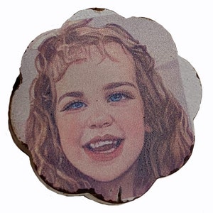 May include: A close-up of a girl's face on a round, white, wooden coaster. The girl has long, brown hair, blue eyes, and a big smile. The coaster has a slightly rough, textured surface.