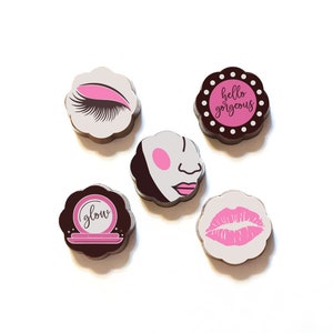 May include: Five chocolate candies with different designs. The designs include a pink eye with eyelashes, a pink face with a nose and lips, a pink compact with the word "glow", a pink lip print, and a pink circle with the text "hello gorgeous".