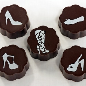 Może przedstawiać: Pięć czekoladowych trufli z różnymi wzorami butów. Każda trufla jest ciemnobrązowa i ma na wierzchu białą sylwetkę buta. Buty obejmują szpilkę, kozak, płaskie buty i dwie różne sandały.