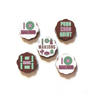 Op de afbeelding: Vijf ronde chocoladebonbons met verschillende ontwerpen. Elke bonbon heeft een witte achtergrond met kleurrijke ontwerpen en tekst. De ontwerpen omvatten een bamboestok, een mahjong-tegel en Chinese karakters. De tekst omvat "Ik hou van Mahjong", "Dot Crack Bam", "Pung Kong Quint" en "Mahjong".