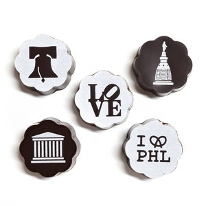 Op de afbeelding: Vijf ronde, zwart-witte chocoladebonbons met verschillende ontwerpen. De ontwerpen omvatten een Liberty Bell, het LOVE-beeld, het Philadelphia City Hall, een pretzel en de woorden "I LOVE PHL".