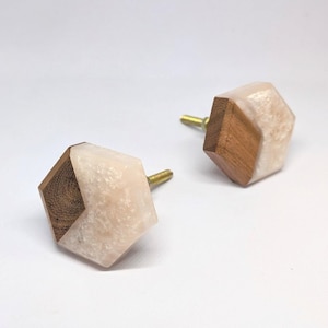 May include: Two decorative cabinet knobs. Each knob features a geometric design with a light pink and white speckled resin section and a dark wood section. The knobs have brass-colored stems.
