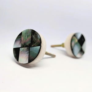 Könnte beinhalten: Zwei dekorative Schrankknöpfe. Jeder Knopf hat eine runde, cremefarbene Basis mit einem Mosaikdesign aus schwarzen, grünen und schillernden Muschelteilen. Die Knöpfe haben einen silberfarbenen Stiel zur Montage.
