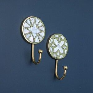 Op de afbeelding: Twee decoratieve wandhaken met een gouden afwerking. Elke haak heeft een cirkelvormig ontwerp met een parelmoer inleg in een geometrisch patroon. De haken zijn op een donkerblauwe muur gemonteerd.