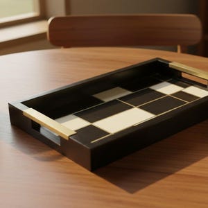 May include: A rectangular black serving tray with brass handles and a chequered black and cream pattern. A glass of iced coffee, coffee beans, and a white teacup and saucer sit on a wooden table.