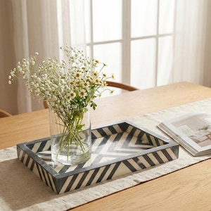 Peut inclure: Un plateau décoratif avec un motif en chevrons noir et blanc contient un vase en verre transparent rempli de fleurs blanches et jaunes. Un magazine ouvert repose sur un chemin de table neutre.