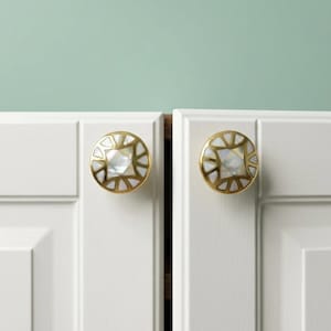 May include: Two circular cabinet knobs featuring a gold-toned metal frame with a geometric pattern. Each knob is inlaid with iridescent white mother-of-pearl segments arranged in a sunburst design, mounted on white cabinet doors with panels.