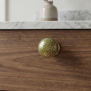 Peut inclure: Un bouton de meuble rond présentant une surface verte brillante avec un motif floral et cachemire doré complexe. Le bouton est monté sur une façade de tiroir en bois brun foncé, avec une base en or métallique reliant le bouton au bois.