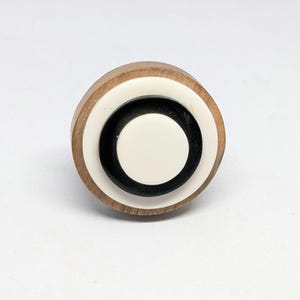 Könnte beinhalten: Ein runder Möbelknopf mit einem äußeren Ring aus naturfarbenem, hellbraunem Holz, einem glatten weißen mittleren Ring und einem massiven schwarzen inneren Ring mit einem kleinen weißen kreisförmigen Zentrum. Das Design besteht aus konzentrischen Kreisen.