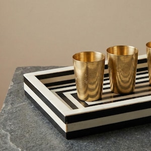 May include: A decorative tray with a black and white geometric pattern holds three gold-colored metal cups. The cups are cylindrical and have a slightly textured surface. The tray is rectangular and sits on a gray stone surface.