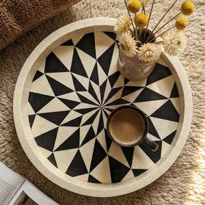 May include: A circular decorative tray featuring a black and white geometric design. It holds a small cup of coffee and a vase of yellow and white dried flowers. The tray rests on a beige rug.