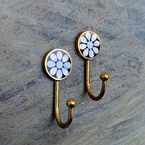 May include: A pair of decorative wall hooks. Each hook has a circular head with a black background and a white flower design. The hooks are gold-coloured, with a curved stem and a small ball at the end. The hooks are designed for hanging items.