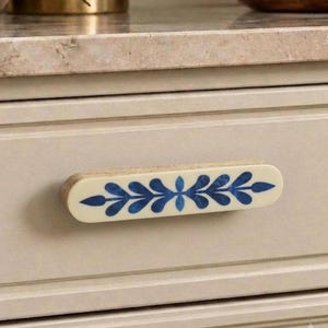 May include: A rectangular drawer pull with rounded ends, featuring a cream-coloured base and a central horizontal blue leaf pattern. The handle is mounted on a light beige drawer front, showcasing a layered wood edge visible on the side profile.