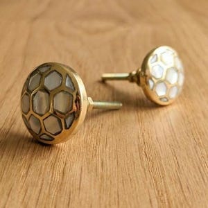 May include: Two decorative cabinet knobs with a gold finish and a honeycomb design. The knobs have a round shape with a mother-of-pearl inlay. The knobs are on a wooden surface.