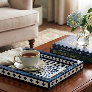 May include: A wooden coffee table with a decorative blue and white tray holding a teacup and saucer, a folded napkin, and a spoon. Two books and a vase of flowers are also on the table. A beige armchair is in the background.