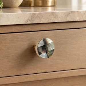 Könnte beinhalten: Ein runder Möbelknopf mit einem Mosaik aus irisierenden, dunkelgrauen und schwarzen Muschelplättchen in geometrischem Muster. Der Knopf hat einen goldfarbenen Metallsockel und ist an einer hellbraunen Holzschublade mit horizontaler Maserung befestigt.
