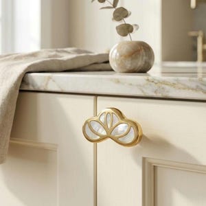 May include: A gold-toned cabinet knob featuring a stylized cloud or lotus shape with inlaid white mother-of-pearl segments. The hardware is mounted on a cream-colored cabinet door, showcasing a metallic frame surrounding the iridescent shell.