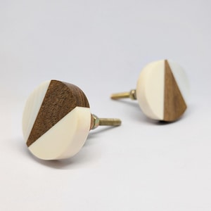 Peut inclure: Deux boutons de meuble décoratifs. Chaque bouton est rond avec un design géométrique. Le design présente un triangle de bois brun foncé, un triangle de matériau blanc cassé et une vis en laiton. Les boutons sont posés sur un fond blanc.