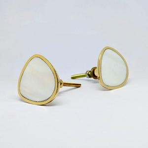 Peut inclure: Deux boutons de meuble décoratifs. Chaque bouton présente une face triangulaire blanche et nacrée avec une bordure en métal doré. Les boutons ont une base dorée et une tige filetée pour le montage. Les boutons sont sur fond blanc.