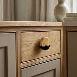 May include: A circular cabinet knob featuring a two-tone design with a natural light brown wood grain top half and a solid black resin bottom half, mounted on a light oak wood drawer front with a visible grain pattern and a frame border.