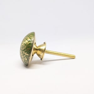 Könnte beinhalten: Ein runder Möbelknopf mit einer grünen Oberfläche und einem goldenen Blumenmuster. Der Knopf verfügt über einen metallisch goldenen Sockel und einen Gewindebolzen aus Metall, der zur Befestigung an Möbeltüren oder Schubladen dient.