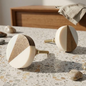 Peut inclure: Deux boutons de meuble décoratifs avec un design géométrique. Chaque bouton présente une combinaison de bois blanc, brun clair et brun foncé. Les boutons ont des vis de couleur laiton pour l'installation. Les boutons sont sur une surface mouchetée.