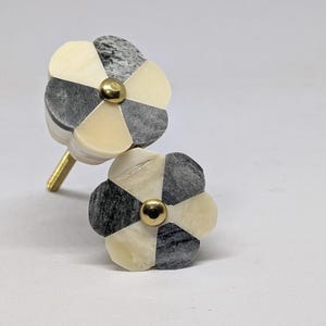 Könnte beinhalten: Zwei blumenförmige Schrankknöpfe mit abwechselnden Segmenten aus cremefarbenem Material und grauem Marmorstein. Jeder Knopf hat einen zentralen goldfarbenen Metallniet und einen goldfarbenen Gewindebolzen an der Rückseite eines der Knöpfe.